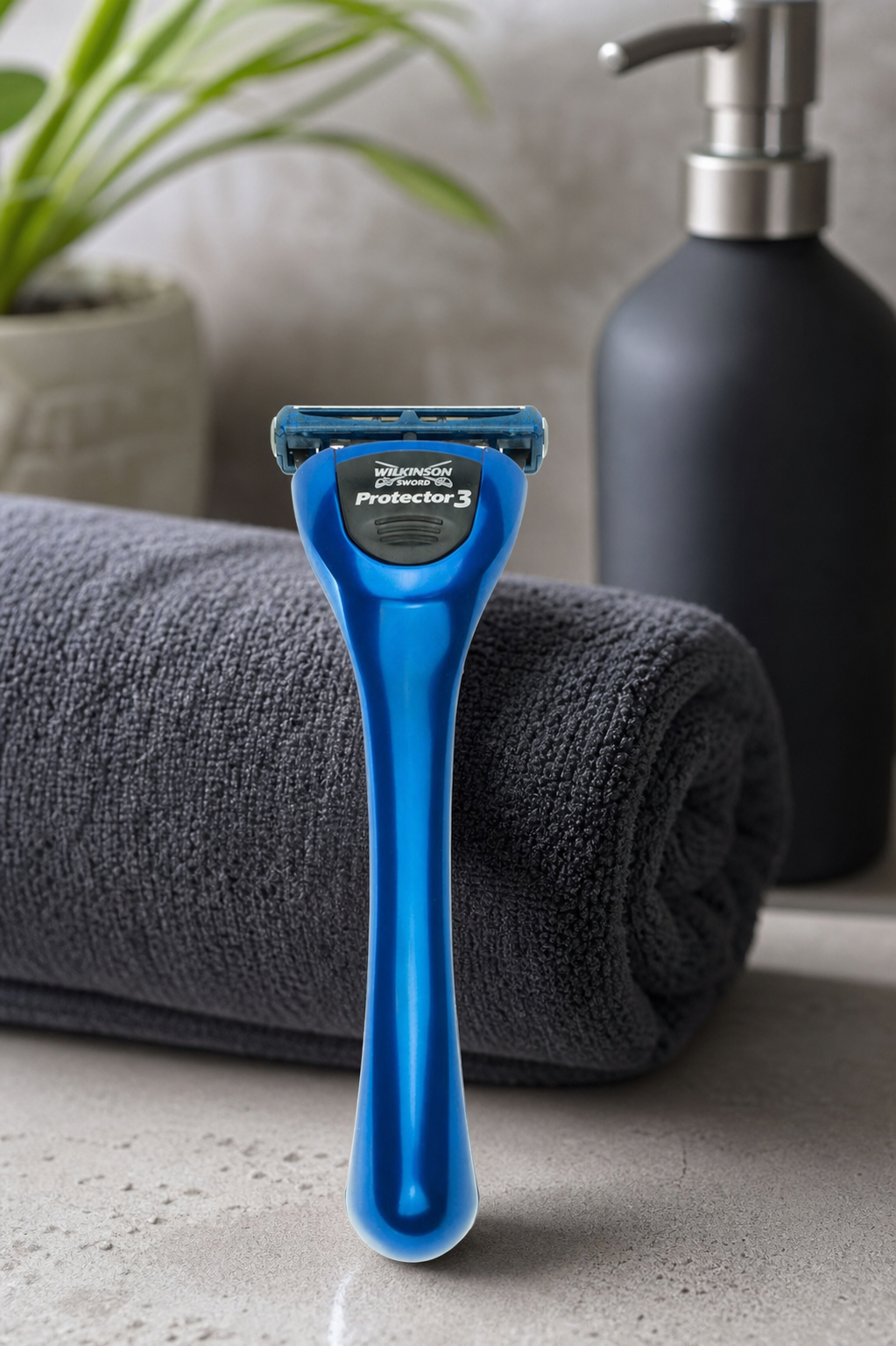 Blauw Wilkinson Sword Protector 3 scheersysteem staand op een badkamerblad naast een opgerolde grijze handdoek en zwarte zeepdispenser